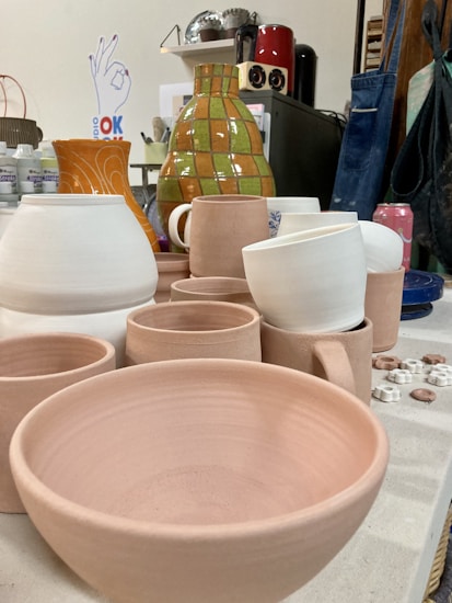 A variety of unfinished clay ceramics, including bowls, cups, and vases, are arranged on a worktable in a pottery studio. Behind them, there are several other pottery tools, a green and orange checkered vase, and various art supplies. A person wearing a blue apron is visible in the background, along with items like bottles, a red canister, and a soda can.