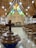 A church sanctuary with wooden ceilings and elongated hanging lights. The room is lined with rows of empty wooden pews. At the front is a large wooden cross on the wall, above an altar covered with red cloths. Behind the cross are vibrantly colored stained glass windows depicting various scenes. In the foreground, there is a brown baptismal font with a prominent cross, and a tiled floor leading up to the altar. The surrounding walls feature banners with the word 'remember' and a drum set is visible to the right side.