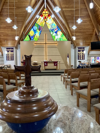 A church sanctuary with wooden ceilings and elongated hanging lights. The room is lined with rows of empty wooden pews. At the front is a large wooden cross on the wall, above an altar covered with red cloths. Behind the cross are vibrantly colored stained glass windows depicting various scenes. In the foreground, there is a brown baptismal font with a prominent cross, and a tiled floor leading up to the altar. The surrounding walls feature banners with the word 'remember' and a drum set is visible to the right side.