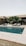 A modern outdoor setting featuring a clear blue swimming pool surrounded by light wooden decking. Several gray lounge chairs are arranged on the deck, with lush green palm trees in the background. The architecture is minimalist with neutral tones.