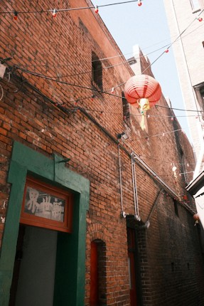 A narrow alleyway with brick walls features a red and white paper lantern suspended by wires. The sunlight creates a warm glow on the bricks. There is also a green-framed door with some text visible above it.