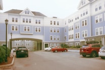 A multi-story residential building displaying a combination of white and light blue siding. Several cars are parked in the foreground, with a driveway leading under an archway in the center. The windows are symmetrically arranged, and there are lampposts lining the sidewalk.