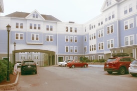 A multi-story residential building displaying a combination of white and light blue siding. Several cars are parked in the foreground, with a driveway leading under an archway in the center. The windows are symmetrically arranged, and there are lampposts lining the sidewalk.
