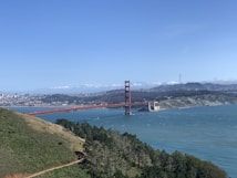 A sweeping landscape view of the iconic Golden Gate Bridge spanning across the water with a backdrop of rolling hills and a distant city skyline. The bridge's distinctive red hue contrasts against the blue sky and ocean. Verdant greenery and trees occupy the foreground, framing the scene with natural beauty.