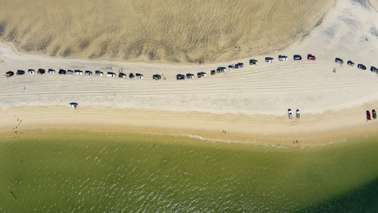 An aerial view of a sandy beach with a long line of parked cars on the upper part of the sand. The beach is bordered by light brown sand with visible tire tracks and a greenish ocean at the bottom. A few people are scattered near the water's edge.