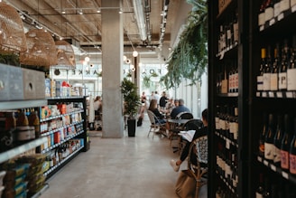 A modern café with an industrial design featuring concrete pillars and exposed ductwork. Shelves filled with wine bottles and assorted packaged goods are prominently displayed along the sides. Patrons are seated at tables, engaging in conversations and enjoying their time. The space is well-lit with hanging woven light fixtures and natural light coming in through large windows. Green plants add a touch of warmth and nature to the setting.