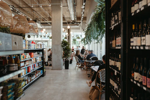 A modern café with an industrial design featuring concrete pillars and exposed ductwork. Shelves filled with wine bottles and assorted packaged goods are prominently displayed along the sides. Patrons are seated at tables, engaging in conversations and enjoying their time. The space is well-lit with hanging woven light fixtures and natural light coming in through large windows. Green plants add a touch of warmth and nature to the setting.