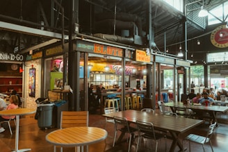 A bustling indoor market scene featuring various food stalls with bright, colorful signage. The tables and chairs are arranged for diners, with groups of people enjoying their meals. The atmosphere feels lively and vibrant, with a focus on Mexican cuisine including tacos and churros.
