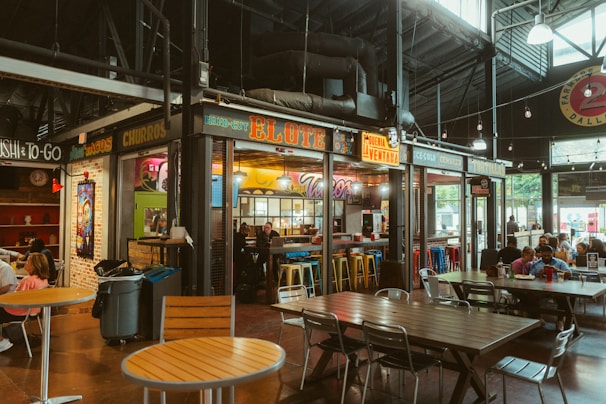 A bustling indoor market scene featuring various food stalls with bright, colorful signage. The tables and chairs are arranged for diners, with groups of people enjoying their meals. The atmosphere feels lively and vibrant, with a focus on Mexican cuisine including tacos and churros.
