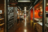 An indoor marketplace features a long, narrow hallway flanked by various eateries and shops. On the left, there is a brick wall with painted signage for a cheese and wine shop. On the right, a sushi restaurant with bright red walls displays Japanese lanterns. Several people are gathered at a counter and walking through the corridor.