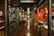 An indoor marketplace features a long, narrow hallway flanked by various eateries and shops. On the left, there is a brick wall with painted signage for a cheese and wine shop. On the right, a sushi restaurant with bright red walls displays Japanese lanterns. Several people are gathered at a counter and walking through the corridor.