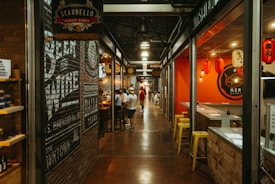 An indoor marketplace features a long, narrow hallway flanked by various eateries and shops. On the left, there is a brick wall with painted signage for a cheese and wine shop. On the right, a sushi restaurant with bright red walls displays Japanese lanterns. Several people are gathered at a counter and walking through the corridor.