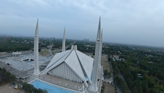 A large, modern mosque with four tall minarets and a striking triangular roof structure, surrounded by landscaped greenery and an urban cityscape in the distance. The sky is overcast, giving a serene, calm ambiance.