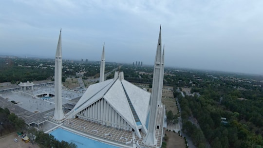 A large, modern mosque with four tall minarets and a striking triangular roof structure, surrounded by landscaped greenery and an urban cityscape in the distance. The sky is overcast, giving a serene, calm ambiance.