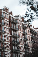 Red brick apartment building featuring white accents and numerous balconies, surrounded by bare tree branches indicative of winter or fall. The architecture is traditional and reminiscent of an older urban style, with several stories and symmetrically arranged windows.