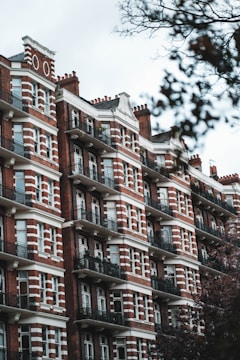Red brick apartment building featuring white accents and numerous balconies, surrounded by bare tree branches indicative of winter or fall. The architecture is traditional and reminiscent of an older urban style, with several stories and symmetrically arranged windows.