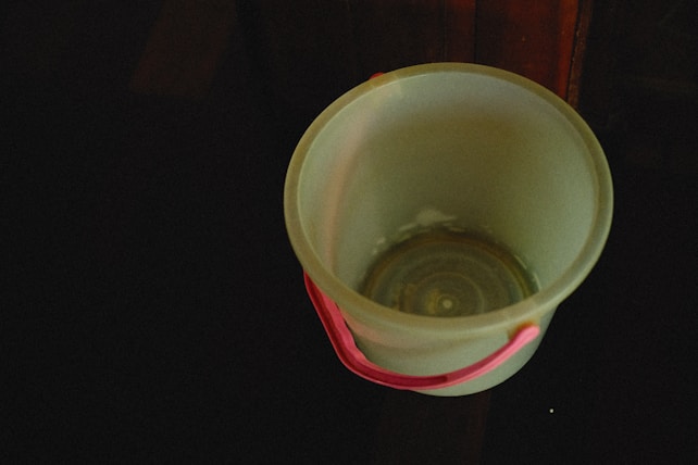 A plastic bucket with a pink handle is placed on a dark surface. The bucket is light-colored and empty.