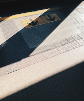 The floor features directional arrows on a yellow strip, surrounded by shadowy and brightly lit patterns cast diagonally across a textured tiled surface.