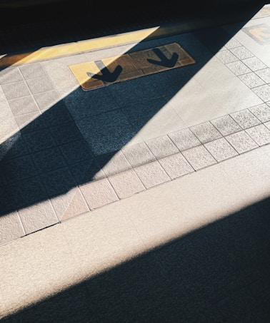 The floor features directional arrows on a yellow strip, surrounded by shadowy and brightly lit patterns cast diagonally across a textured tiled surface.