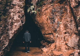 A person wearing a dark jacket stands at the entrance of a cave. The cave is surrounded by large, rugged rock formations with patches of moss and small plants growing on the surface. The atmosphere is dim and mysterious, suggesting an exploration or adventure.