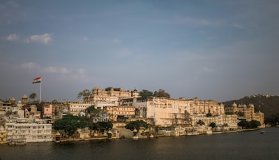 A sprawling palace complex sits majestically on the shores of a serene lake, featuring traditional architecture with intricate details and a large Indian flag flying prominently. The buildings are a mix of cream and light brown colors, set against a backdrop of lush greenery and distant hills under a partially cloudy sky.