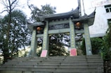 A traditional stone gate with ornate Chinese architectural elements stands surrounded by trees and greenery. Red lanterns and intricate golden Chinese characters adorn the gate. The structure includes curved, layered roofs typical of classical Chinese buildings.