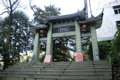 A traditional stone gate with ornate Chinese architectural elements stands surrounded by trees and greenery. Red lanterns and intricate golden Chinese characters adorn the gate. The structure includes curved, layered roofs typical of classical Chinese buildings.