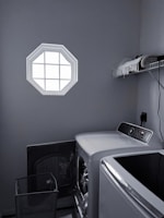 A laundry room with a washer and dryer set is visible. The washer door is open, revealing some clothes inside. There is a small octagonal window on the wall allowing light into the room. A shelf above the appliances holds folded towels and a few other items. The room is mostly gray with dim lighting.