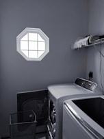 A laundry room with a washer and dryer set is visible. The washer door is open, revealing some clothes inside. There is a small octagonal window on the wall allowing light into the room. A shelf above the appliances holds folded towels and a few other items. The room is mostly gray with dim lighting.