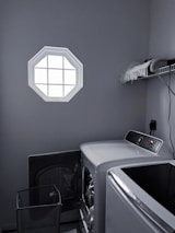 A laundry room with a washer and dryer set is visible. The washer door is open, revealing some clothes inside. There is a small octagonal window on the wall allowing light into the room. A shelf above the appliances holds folded towels and a few other items. The room is mostly gray with dim lighting.