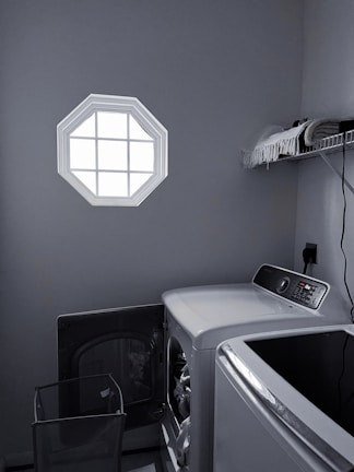 A laundry room with a washer and dryer set is visible. The washer door is open, revealing some clothes inside. There is a small octagonal window on the wall allowing light into the room. A shelf above the appliances holds folded towels and a few other items. The room is mostly gray with dim lighting.