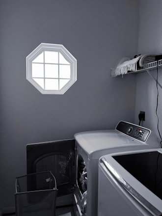 A laundry room with a washer and dryer set is visible. The washer door is open, revealing some clothes inside. There is a small octagonal window on the wall allowing light into the room. A shelf above the appliances holds folded towels and a few other items. The room is mostly gray with dim lighting.