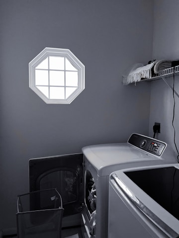 A laundry room with a washer and dryer set is visible. The washer door is open, revealing some clothes inside. There is a small octagonal window on the wall allowing light into the room. A shelf above the appliances holds folded towels and a few other items. The room is mostly gray with dim lighting.