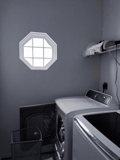 A laundry room with a washer and dryer set is visible. The washer door is open, revealing some clothes inside. There is a small octagonal window on the wall allowing light into the room. A shelf above the appliances holds folded towels and a few other items. The room is mostly gray with dim lighting.