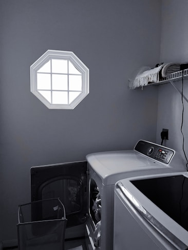 A laundry room with a washer and dryer set is visible. The washer door is open, revealing some clothes inside. There is a small octagonal window on the wall allowing light into the room. A shelf above the appliances holds folded towels and a few other items. The room is mostly gray with dim lighting.