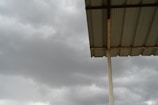 A corrugated metal roof supported by a vertical metal beam, set against a backdrop of overcast, cloudy skies. The sky appears dark grey, suggesting an approaching storm or rain.