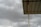 A corrugated metal roof supported by a vertical metal beam, set against a backdrop of overcast, cloudy skies. The sky appears dark grey, suggesting an approaching storm or rain.