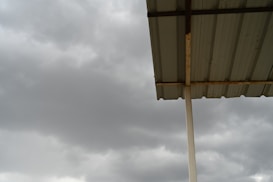 A corrugated metal roof supported by a vertical metal beam, set against a backdrop of overcast, cloudy skies. The sky appears dark grey, suggesting an approaching storm or rain.