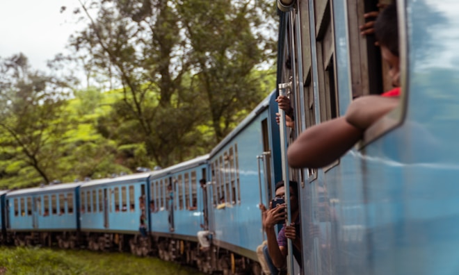 A blue train is winding through a lush green landscape. Passengers are leaning out of the windows, taking photos and enjoying the scenery. Trees and greenery surround the railway track, contributing to a serene environment. The train appears to be moving at a slow pace, given the relaxed demeanor of the passengers.