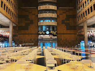 A large indoor space featuring rows of empty white folding chairs arranged in a symmetrical pattern on a tiled floor. The area appears to be an atrium in a public building, with high ceilings and modern architectural elements. A prominent brown and black wall is in the background, adorned with clocks and decorative gears. The space is well-lit, with blue and white lighting highlighting different sections.