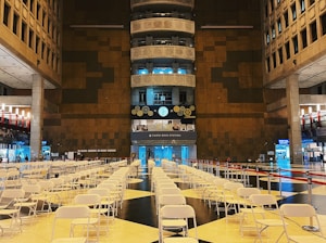 A large indoor space featuring rows of empty white folding chairs arranged in a symmetrical pattern on a tiled floor. The area appears to be an atrium in a public building, with high ceilings and modern architectural elements. A prominent brown and black wall is in the background, adorned with clocks and decorative gears. The space is well-lit, with blue and white lighting highlighting different sections.