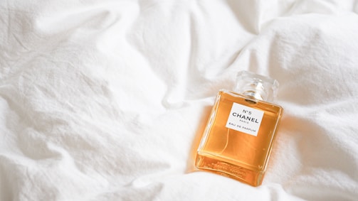 A bottle of Chanel No. 5 perfume rests on a soft, white quilt, highlighting its elegant design and golden liquid.