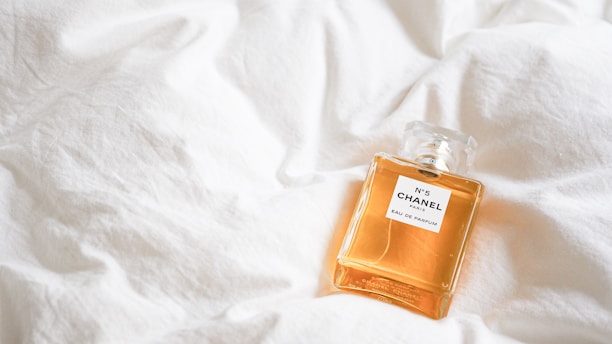 A bottle of Chanel No. 5 perfume rests on a soft, white quilt, highlighting its elegant design and golden liquid.