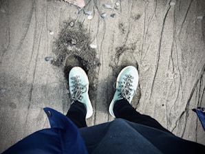 A pair of light blue sneakers with laces stand on a sandy surface, with footprints slightly visible in the sand. The sand is textured with various small stones scattered around. The person appears to be wearing black pants and a blue jacket.