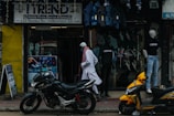 A street scene featuring a shop with a sign reading 'Trend' offering mobile phone service and sales. Mannequins displaying clothing are visible in the storefront. Two motorcycles, one black and one yellow, are parked outside on the sidewalk. A person wearing traditional attire walks past the store.