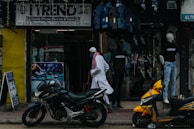 A street scene featuring a shop with a sign reading 'Trend' offering mobile phone service and sales. Mannequins displaying clothing are visible in the storefront. Two motorcycles, one black and one yellow, are parked outside on the sidewalk. A person wearing traditional attire walks past the store.