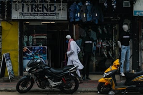 A street scene featuring a shop with a sign reading 'Trend' offering mobile phone service and sales. Mannequins displaying clothing are visible in the storefront. Two motorcycles, one black and one yellow, are parked outside on the sidewalk. A person wearing traditional attire walks past the store.