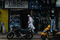 A street scene featuring a shop with a sign reading 'Trend' offering mobile phone service and sales. Mannequins displaying clothing are visible in the storefront. Two motorcycles, one black and one yellow, are parked outside on the sidewalk. A person wearing traditional attire walks past the store.