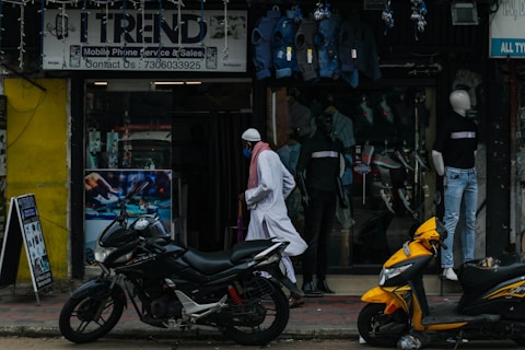 A street scene featuring a shop with a sign reading 'Trend' offering mobile phone service and sales. Mannequins displaying clothing are visible in the storefront. Two motorcycles, one black and one yellow, are parked outside on the sidewalk. A person wearing traditional attire walks past the store.