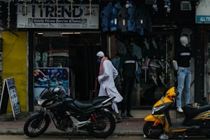 A street scene featuring a shop with a sign reading 'Trend' offering mobile phone service and sales. Mannequins displaying clothing are visible in the storefront. Two motorcycles, one black and one yellow, are parked outside on the sidewalk. A person wearing traditional attire walks past the store.